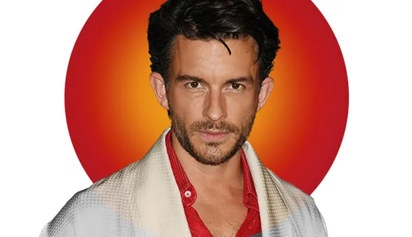 Portrait of actor Jonathan Bailey with dark wavy hair, wearing a red shirt under a white and gray layered outfit. A vibrant red and orange circular background highlights his face. Featured in Madame Tussauds' 'Hot 100' list.